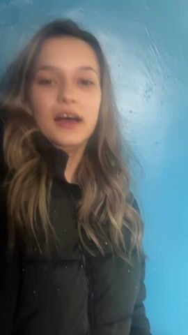 The video features a young woman with long, wavy brown hair, standing against a bright blue wall. She is wearing a black, puffy jacket. She begins by talking, then smiles broadly.
