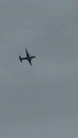 A twin-engine turboprop aircraft flies across a cloudy sky. The aircraft is seen from below, banking slightly to its right. The underside of the aircraft is visible, along with its wings, engines, and tail. A dark angled structure, possibly the edge of a roof or building, is visible in the lower right corner of the frame, obscuring part of the sky. The overall lighting suggests an overcast day.