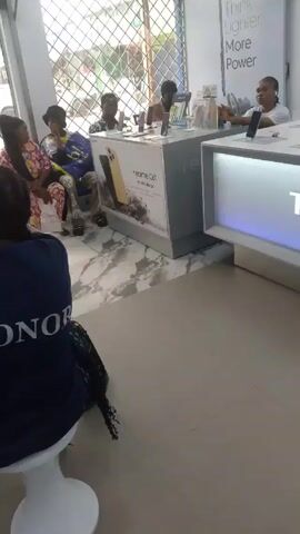 The video shows the interior of a mobile phone store.  Several customers are seated and standing, observing and interacting with the sales staff. One woman wearing a blue shirt with "HONOR" printed on the back sits on a white stool facing a display table.  A person stands near the entrance, which is open to a street scene, as people and vehicles pass by. There are display counters featuring Realme and Honor products. The store has a modern appearance, with tiled floors and bright lighting.