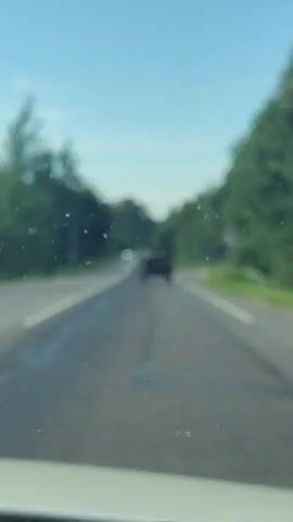 The video shows a road in Agalatovo, Russia, on a sunny day. The road is paved and has two lanes in each direction, separated by a white line. Trees line both sides of the road, and there are power lines overhead. In the distance, a convoy of vehicles is visible, including a military-style jeep in the foreground. The sky is clear and blue, suggesting good weather. The overall mood appears to be calm and peaceful, with no immediate signs of distress or urgency. The scene is set in a rural or suburban area, with the dense foliage indicating a natural or semi-natural environment. There are no people visibly identifiable in the video. The video is captured from a moving vehicle, likely a car, as evidenced by the perspective and the view of the dashboard in the foreground.
