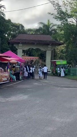 The video shows an entrance gate with a traditional tiled roof leading into a park or temple grounds in Sri Lanka. People, mostly dressed in white, are walking through the gate. Several vendors with food stalls are positioned on either side of the entrance, with a bright pink tent on the left. A green tuk-tuk is parked on the right, and a beige tuk-tuk sits off to the side on the grass. A large stone with carved Sinhalese writing is visible to the right of the gate. The sky is cloudy and the scene is surrounded by trees.
