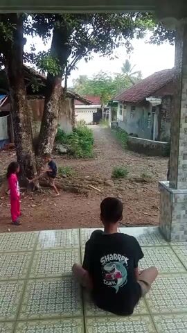 A boy in a dark t-shirt with a shark graphic sits on a tiled porch, facing away from the camera. In the background, a young girl in a bright pink outfit and a young boy in a blue shirt and brown shorts are playing. The girl is standing near a tree, while the boy is slightly further away, kicking a yellow ball. The environment appears to be a yard with a dirt path, some trees, and a few rustic buildings in the background, one covered with a blue tarp.
