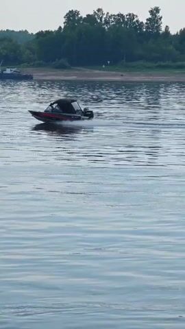 A motorboat speeds across a body of water, leaving a wake. The boat is red and black and has a black canopy. The water is calm and reflects the sky. In the background, there is a shoreline with trees and a few buildings. A tugboat is docked near the shore. The sky is overcast.
