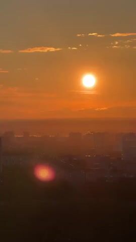A breathtaking sunset unfolds over a vast city skyline. The bright, round sun hangs prominently in the upper center of the frame, casting a warm, orange-red glow across the entire sky. A few wispy clouds, tinged with the same fiery hues, are scattered above and to the left of the sun. Below, a sprawling urban landscape stretches to the horizon, with countless buildings silhouetted against the bright sky. A soft haze blankets the distant parts of the city, enhancing the atmospheric effect of the sunset. A prominent lens flare, appearing as a bright pink and orange orb, is visible in the lower central part of the video, caused by the direct light of the sun hitting the camera lens. The scene captures a tranquil and picturesque end to the day.