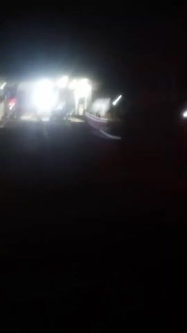 The video appears to be shot from a moving vehicle at night. In the foreground, a blurred red light is visible, possibly a taillight. In the background, a bright, illuminated building or structure is seen. The building is lit with a strong, white light, and there are hints of other lights and possibly people or vehicles around it, but they are all heavily blurred due to the low light conditions and movement. The overall impression is of a somewhat desolate or rural setting at night with a focal point of activity around the illuminated building.