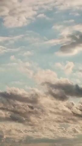 The video is a close-up shot of the ocean and the sky. The sky is filled with clouds, some of which are dark and heavy, while others are light and fluffy. The sun is not visible, but its light is filtering through the clouds, creating a soft, diffused glow. The ocean is dark blue and calm, with gentle waves lapping at the shore. The video is very peaceful and serene.