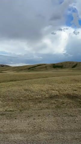 This video captures a panoramic view of a vast, open landscape in Bakty, Kazakhstan. The scene is dominated by rolling hills covered in dry, golden grass, indicative of a late summer or autumn season. In the foreground, a gravel road runs horizontally across the frame, suggesting this is a rural or undeveloped area.

The sky is dramatic and dynamic, filled with a mixture of dark, heavy storm clouds and patches of bright blue sky where sunlight breaks through. This creates a sense of changing weather, with potential for rain or a storm approaching or passing. The lighting is varied, with some areas of the landscape brightly illuminated and others cast in shadow.

The overall mood is one of natural grandeur and perhaps a touch of isolation, given the expansive and seemingly uninhabited terrain. The rolling hills stretch towards the horizon, where a faint line of mountains can be seen in the distance. There are no people or man-made objects, apart from the road, visible in the frame, emphasizing the natural and untamed character of the location. The time of day appears to be afternoon or late afternoon, based on the angle of the light. There is no visible text in the video.
