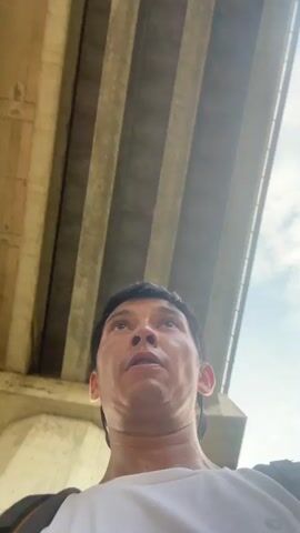 The video shows a low-angle, first-person perspective shot of a man looking upwards. He is standing under a large concrete structure, possibly a bridge, with several parallel beams visible overhead. Sunlight filters through the gaps between the beams. The man's face fills the lower portion of the frame. He appears to be in motion, as his head bobs and his facial expressions change. In the first few seconds, he looks up with a neutral expression, then his face moves closer to the camera and he makes a surprised or disgusted face, with his mouth in a pout and his eyebrows raised. Green foliage is visible to the left of the frame at one point, suggesting an outdoor, possibly vegetated, environment.