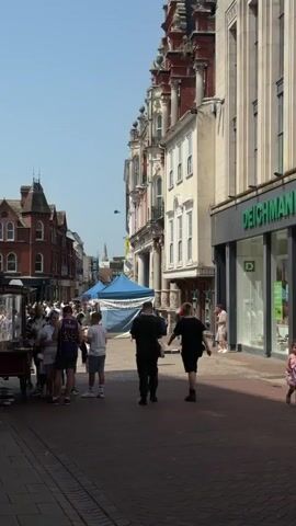 The video shows a pedestrian street scene on a sunny day. Several people walk along the brick-paved street, some of whom are carrying shopping bags. The buildings that line the street vary in architectural style, ranging from old-fashioned brick structures to more modern designs. One building has a "Deichmann" store on the ground floor. Several tents are set up along the street, creating a market-like atmosphere. A busker is standing near one of the storefronts, performing for the people walking by. Overall, the scene depicts a bustling commercial area.