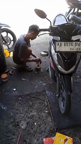 In a medium shot, a person is working on a black Honda scooter inside what appears to be an automotive repair shop. The person, a man with short dark hair, is squatting near the front of the scooter. He is wearing a dark t-shirt and dark sandals. He is holding a white bottle and appears to be applying some kind of liquid to a component near the scooter's handlebars. The scooter has a license plate that reads "AE 6576 DW" and an expiration date of "04-29". The shop floor is dark and dirty with scattered tools, parts, and containers. Parts of other motorcycles are visible in the background, partially obscured. There is a can of yellow liquid on the left and a small red package and a small yellow package on the right on the ground.