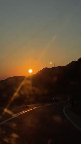 The video shows a scenic sunset over a winding road. The sun is a bright orange orb low in the sky, casting a warm glow over the landscape. Silhouetted mountains form the backdrop, with a few trees visible on the right side. The road itself is dark asphalt with a white dividing line, and a few cars are driving on it. The sky transitions from a light orange near the sun to a dusky blue at the top. There are some light flares and bokeh effects present, suggesting the video was filmed through a lens that is slightly dirty or has some obstruction. The overall mood is peaceful and serene.
