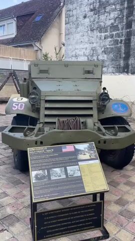 The video shows a static display of two military vehicles. In the foreground, on the right, is a green halftrack vehicle with the number 50 on its front fender and an anchor insignia. A sign in front of it provides information about the vehicle in multiple languages. To the left of the halftrack is a teal-colored tank. The video pans from the halftrack to the tank, offering different views of both vehicles. The environment appears to be an outdoor display area, possibly a museum or memorial, with paved walkways and some greenery, including palm trees, in the background. The sky is visible with clouds. There are no people interacting with the vehicles.