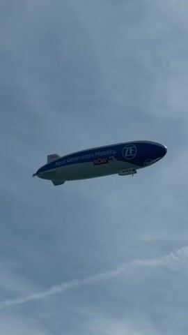 A blimp is seen floating in the sky against a cloudy blue sky. The blimp is dark blue with white lettering that reads "Next Generation Mobility." In the center of the blimp, there is a white circle with blue lettering that reads "ZF". Below the "ZF" logo, the word "NOW" is displayed in red lettering. The blimp appears to be in motion.