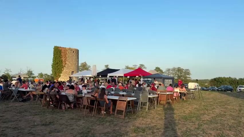 This is a wide shot of an outdoor gathering. A large group of people are seated at tables set up on a grassy field. Tents of various colors are erected behind the tables, providing shade. To the left, there is a large inflatable structure, possibly a bouncy castle. In the background, a round stone tower covered in ivy stands prominently. A man in a red shirt and blue shorts is standing in the foreground, facing the camera and holding an object. The sky is clear and blue, suggesting it is daytime. Several cars are parked in the background.
