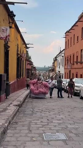 The video shows a cobblestone street in a town or city with colorful buildings on either side.  A young woman in a large pink dress is walking down the street, surrounded by several other people. Some of the people appear to be helping her carry the dress, while others are walking alongside her. There are cars parked on one side of the street. In the distance, the cityscape is visible. The sky is blue with some clouds. The overall atmosphere appears festive and celebratory.