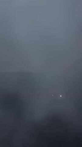 The video shows a foggy night scene with a dark, angled structure on the left, likely a building or roof. In the distance, faint lights are visible, possibly from other buildings or streetlights, struggling to penetrate the thick fog. The fog itself appears to be swirling and moving, especially in the lower part of the frame, creating an ethereal and slightly mysterious atmosphere. The overall impression is one of low visibility and a quiet, perhaps somewhat isolated, nighttime environment in Qaskeleñ, Kazakhstan. The time of day is clearly night due to the darkness and artificial lights. The weather condition is heavy fog. There is no visible text in the video.