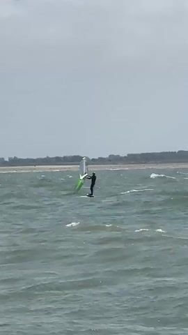 A person is windsurfing on a choppy body of water under a cloudy sky. The windsurfer is wearing dark clothing and is standing on a board. They are holding onto a sail that is predominantly white with a green section. In the background, a distant landmass with trees is visible along the horizon. The water is a murky green-gray, and small waves are breaking. The overall impression is a dynamic scene of outdoor recreation in potentially overcast weather.