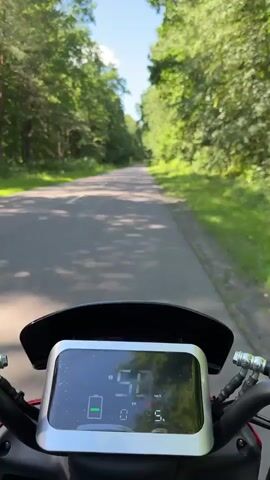 A first-person perspective video shows a digital dashboard of an electric scooter or motorcycle as it travels down a paved road surrounded by trees. The dashboard displays a battery icon, speed in kilometers per hour, and a trip odometer. The road has a white line marking on the left side and is bordered by lush green trees on both sides. The sky is visible above the trees and appears to be clear and blue. The scooter is moving forward at a consistent speed.