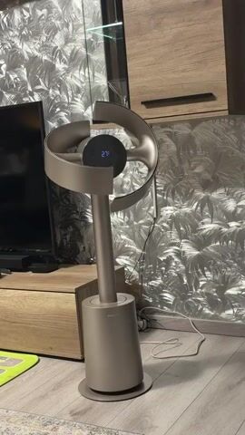 A fan with a digital display showing 27 degrees Celsius is positioned in a room. The fan has a unique, modern design with a circular head and a slender stand. A power cord is plugged into the base of the fan and extends across the floor. The background of the room features a patterned wallpaper and wooden furniture. The lighting appears to be interior lighting. The fan is stationary and no people are visible in the frame.