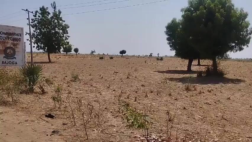 The video opens with a daytime shot of a sign that reads "Welcome to Gombe State Polytechnic, Bajoga." The surrounding environment is a dry, brown field with scattered vegetation. The camera then moves, suggesting it is mounted on a vehicle, showing a similar dry landscape with trees and power lines. A paved road comes into view, leading toward the horizon with a red truck visible in the distance.