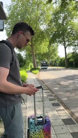 A man with a suitcase stands at a bus stop, holding a phone in his hands. He's wearing sunglasses and earbuds. The suitcase, positioned beside him, has a colorful, animal print design. A bus approaches in the distance, displaying the number "147" and the word "Station". The surrounding environment includes trees and greenery alongside the road.