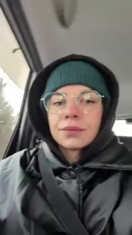 A person with light skin, wearing a green beanie, round glasses, a black hooded jacket, and a seatbelt, is in a car. They hold up a handheld computer with a screen, then put it down. The view is from the driver's perspective. The car appears to be moving, with trees visible through the window.  The lighting is natural and suggests an overcast day.