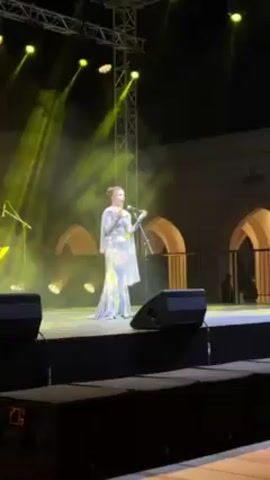 A woman stands on a stage and sings into a microphone. She is wearing a long, pastel gown with intricate detailing, and a sheer cape. She appears to be singing with great emotion, gesturing with her hands as she performs. The stage is lit with bright yellow spotlights, and there are speakers in the foreground. Behind her is a building with arched windows, and a lighting rig is visible above the stage. The environment suggests an outdoor performance or concert setting.