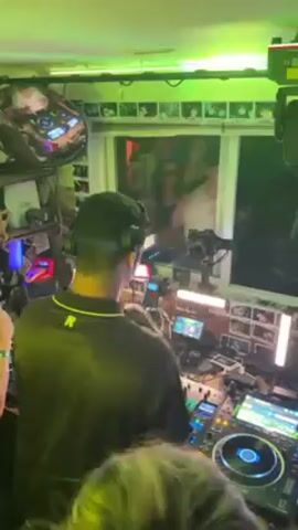 The video shows a DJ at a party. He is standing behind a DJ setup, wearing headphones and a black shirt with a yellow emblem. There are other people in the room, mostly visible from the back, who are dancing and raising their hands in the air. The room is lit with green lights. Behind the DJ there is a window and a wall covered with photos. There are screens and equipment visible on the DJ's table and hanging on the walls.