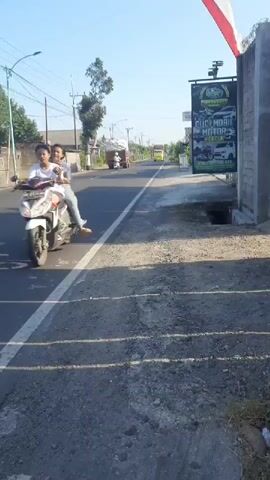 A video taken from a roadside perspective shows a street scene on a sunny day. In the foreground, there is a rough, gravelly shoulder with shadows cast from an unseen object. The road is paved with a white line marking the edge of the traffic lane. Several vehicles are visible on the road, including a motorcycle traveling away from the camera, a dark-colored SUV, and two trucks. The truck in the foreground is yellow and appears to be a large cargo truck. On the right side of the road, there is a gray wall and a tall, dark green sign advertising a car and motorcycle wash. The sign features images of vehicles. A red and white flag is visible at the top right of the frame. Trees and utility poles are present in the background. The sky is clear and blue.