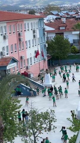 The video shows a schoolyard filled with children in green uniforms. They are spread throughout the courtyard, with some running, some standing, and some interacting with each other. A light post stands in the foreground. The school building is a pink and blue structure with multiple air conditioning units. Two Turkish flags are visible. In the background, there are residential buildings with red tile roofs and a mountainous landscape. The camera is positioned at an elevated angle.