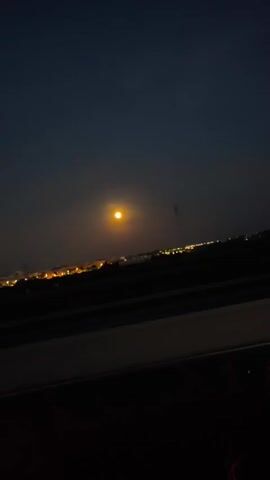 The video shows a night scene with a full moon over a city skyline. The moon appears bright orange, contrasting against the dark blue sky. Below, the city lights are visible as a row of orange and white dots along the horizon. A roadway is in the foreground, with dark asphalt and a bright white concrete barrier. There's a flickering reddish light visible at the bottom right corner of the frame, possibly from another vehicle. Occasional streaks flash across the image.  The camera appears to be moving, potentially from a vehicle traveling along the road. The overall environment is dark and illuminated mainly by the moon and city lights.