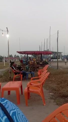 A group of people are seated in orange plastic chairs around orange plastic tables. In the background, there is a vendor cart with a red canopy, displaying various items. A volleyball net is visible in the distance. The sky is overcast.