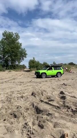 The video shows a bright green SUV driving on a sandy track. The SUV has a roof rack with an awning. In the background, there are trees and a cloudy blue sky. A few people are visible in the distance to the right of the vehicle. The camera is positioned low, looking up at the car and the sky. The sand shows tire tracks from previous vehicles.