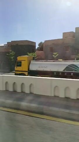 The video, taken from a moving vehicle, presents a view of a street scene on a sunny day. A white, arched-wall barrier is visible in the immediate foreground. A yellow truck passes by on the road beside the barrier.  Beyond the road, there is a low, multi-level building with a somewhat aged appearance. Further along, the view opens up to reveal a modern-style building and a mosque with minarets in the distance. An advertising board with the number 75 displayed can also be seen. There are cars and trees visible along the road. The sky is blue with a clear view of the sun.