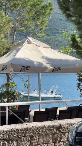 A bright, sunny outdoor scene with a large beige umbrella providing shade over a seating area. The umbrella has several logos on it. In the background, the blue water of a bay or lake is visible, with a white inflatable boat with a console and seating moving on the water. Mountains covered in green foliage rise in the distance. In the foreground, there are several dark chairs and a stone wall. The leaves of trees frame the top of the image.