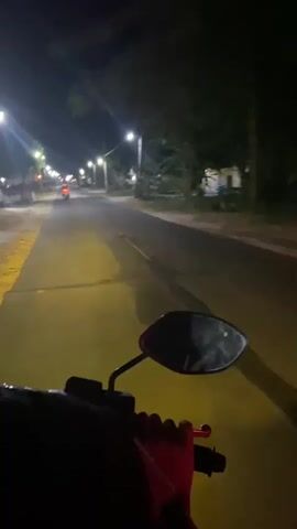 The video shows a first-person perspective of someone riding a motorcycle at night on a paved road. The road is lit by streetlights, which cast a yellow glow. Trees and buildings line the sides of the road. A motorcycle is visible in the distance in front of the rider. The rider's hand is visible on the handlebar. The camera is pointed forward, capturing the road ahead.
