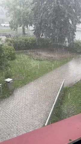 This video captures a hailstorm in Tábor, Czechia. The scene depicts a residential area with a paved walkway, grassy lawns, and trees. A white car is visible in the background, along with a building. The weather is characterized by a heavy downpour of hail, with individual hailstones appearing as white streaks against the green foliage and gray pavement. The sky is overcast, suggesting it is daytime but without direct sunlight. The overall mood is one of a dramatic natural event, with the intensity of the hail creating a dynamic visual. No text is visible in the video.