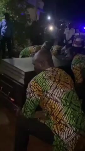At night, a group of men wearing matching brightly-colored shirts are lined up behind a dark-colored coffin. Other people are gathered around them, some holding phones with their lights on to illuminate the scene. The men appear to be lifting or moving the coffin, while the surrounding crowd watches. The setting is outdoors, near a building and some greenery.
