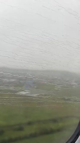 The video pans across a window with raindrops on it, obscuring the view of the landscape below. The landscape appears to be a rural area with green fields and scattered buildings. The sky is overcast and appears to be raining. The video is shot from a moving vehicle, likely an airplane, as indicated by the curved edge of the window.