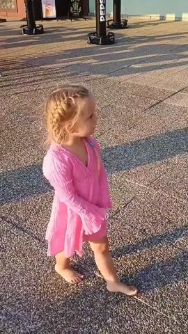 A young blonde child wearing a pink dress stands on a textured outdoor surface, possibly a walkway. The child has braided hair and is looking to the right of the frame. The sun is casting long shadows, suggesting it is either early morning or late afternoon. In the background, blurred figures of adults are visible, and there are some upright objects, possibly poles or stanchions. The child shifts their weight and moves their arms slightly.