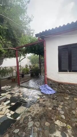 Rain is falling on a patio area outside a house. The patio is made of cobblestones and some broken tiles. A blue tarp is lying on the ground near the house. There is a red metal pergola with some plants growing on it. Trees and more plants are visible in the background. The sky is overcast and appears to be heavy with rain.