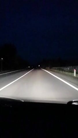 This is a first-person perspective video of driving on a dark, paved road. The headlights of the vehicle illuminate the road ahead, with white lines marking the lanes. The sky is a deep, dark blue, indicating it is nighttime. Trees and vegetation line the sides of the road. There are occasional road signs visible. The video is static and does not show any specific actions or interactions.