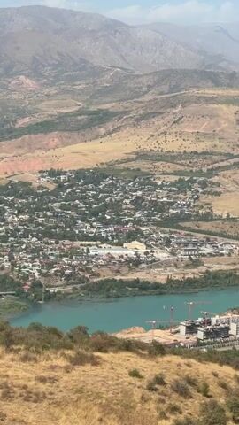The video opens with a panoramic view of a vast, arid landscape dominated by dry, brown hills and mountains under a hazy sky. In the mid-ground, a town or village is nestled in a valley, characterized by numerous small buildings with red and brown roofs, interspersed with patches of green trees. A vibrant turquoise body of water, possibly a lake or reservoir, runs through the valley, with construction cranes and partially built structures visible along its banks. The foreground is occupied by dry, golden grass and low-lying green shrubs. The camera then shifts to show the interior of a modern cable car cabin. Two individuals, a man and a child, are seated inside. The man, wearing a light-colored traditional garment, is looking out the window, while the child, also in similar attire, appears to be resting. The cable car's window displays the reflection of the landscape outside and the text "ChinoRkent" and "021". The video briefly returns to the sweeping landscape view, similar to the opening shot, before ending.