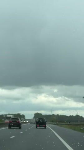 The video shows a driver's perspective while driving on a highway. The weather appears overcast, with a grey sky and rain visible on the windshield. Several cars and a truck are visible on the road ahead. Wind turbines can be seen in the distance on the right side of the road, beyond a line of trees. The highway has multiple lanes, marked by white lines.