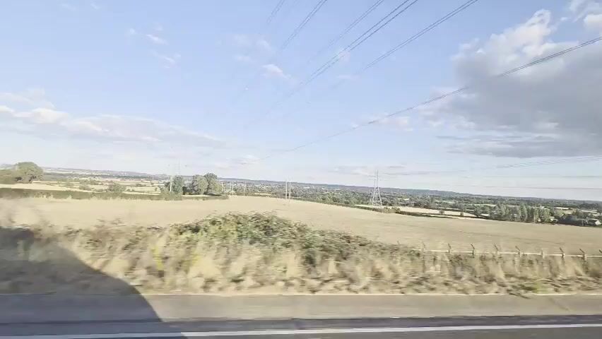 The video shows a landscape seen from a moving vehicle, likely a car or train. The perspective is slightly tilted and from a side window. A dry, grassy field is in the foreground, separated by a fence. Beyond the field, there are more fields, some trees, and in the distance, buildings that appear to be part of a town or village. Power lines and pylons are visible across the landscape, suggesting it is a rural or semi-rural area. The sky is blue with scattered white clouds, and the lighting indicates it is daytime, possibly late afternoon due to the soft light. There are no people or animals visible in the frame.