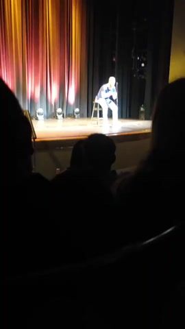 A man with red hair and a beard is sitting on a stool on a stage, holding a microphone. He is wearing a blue t-shirt and light-colored jeans. The stage has a backdrop of red and yellow curtains and stage lights. The audience is visible in the foreground, silhouetted in the dark. The man appears to be speaking or performing.
