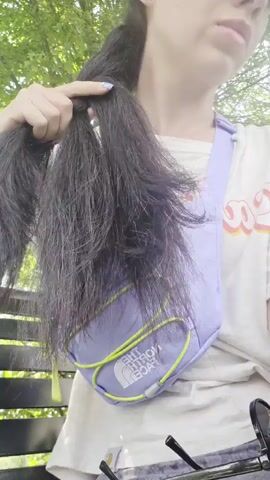 A young woman with dark hair is sitting outside on a bench, braiding her hair. She is wearing a light-colored t-shirt, a purple fanny pack, and a watch. Her hair is partially braided into two sections. The background is blurred, showing green foliage.