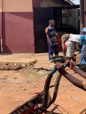 Here is a concise description of the video:

The video depicts a group of people, mainly women, gathered outdoors in what appears to be a communal or market setting. They are standing around low platforms or raised areas, working with bowls and objects that seem to be fish or some other type of food product. Several people are actively engaged in cleaning or preparing the items, while others observe or sit nearby. The environment is dusty and unpaved, and there is a building or structure in the background with dark windows. The people are dressed in a variety of colorful clothing, suggesting a diverse group.