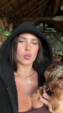 The video shows a woman with dark hair and a black hoodie holding a small Yorkshire Terrier. She is puckering her lips as if to kiss the dog, and the dog is looking towards the camera. The background appears to be an indoor space with wooden beams and furniture. The lighting is dim.