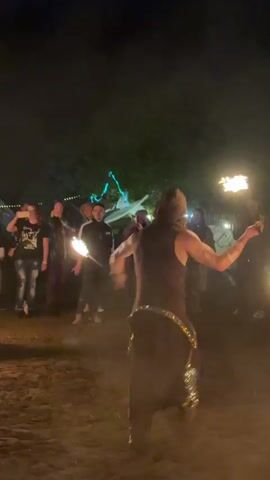 Two fire performers, a woman in a light-colored dress and a man in dark attire, dance with flaming props on a dirt ground at night. The woman wields a fire staff, while the man spins two smaller fire props. Several small fire pots burn on the ground near them. A crowd of onlookers is visible in the background, alongside dimly lit tents and structures under the dark sky.